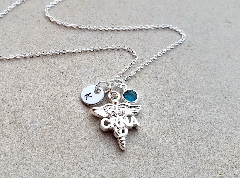 May include: Silver necklace with a charm of a medical caduceus with the letters "CRNA" on it. There is also a small silver charm with the letter "K" on it and a teal blue gemstone.