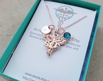 May include: Rose gold necklace with a medical caduceus charm, a small round charm with a letter 'C', and a teal blue gemstone. The necklace is in a teal blue gift box with a white card that says "Amula Designs Etsy Healthcare themed jewelry on pins and www.amula.etsy.com http://amula.etsy.com"
