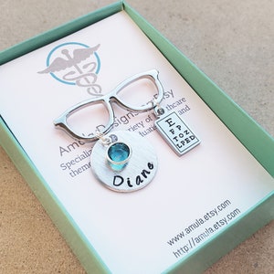May include: A silver charm necklace with a pair of glasses, a small blue gemstone, and a personalized name tag that says "Diane". The charm also includes a small eye chart with the letters "E, FP, TOZ, LPED". The necklace is in a green gift box with the text "www.amula.etsy.com" and "http://amula.etsy.com".