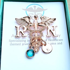 May include: Rose gold-toned 'RN' pin with a caduceus symbol and a blue crystal charm. The pin is on a white card with the text 'Specializing in themed jewelry and pins and of Healthcare'.
