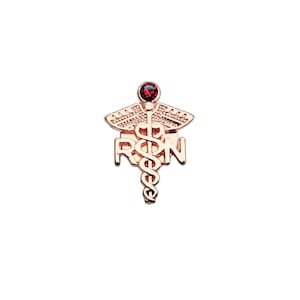Puede incluir: Un broche de metal en tono oro rosa que presenta un s&iacute;mbolo de caduceo con las letras RPN integradas en el dise&ntilde;o. La parte superior del caduceo est&aacute; adornada con una peque&ntilde;a piedra preciosa redonda de color rojo oscuro engastada.