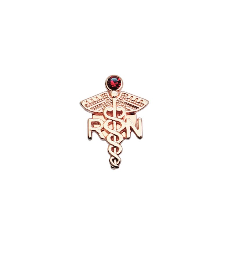 Puede incluir: Un broche de metal en tono oro rosa que presenta un s&iacute;mbolo de caduceo con las letras RPN integradas en el dise&ntilde;o. La parte superior del caduceo est&aacute; adornada con una peque&ntilde;a piedra preciosa redonda de color rojo oscuro engastada.