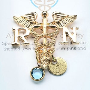 Puede incluir: Pin de caduceo médico dorado con un dije de cristal azul y un dije de oro con la letra J. El pin presenta las letras RN a cada lado del caduceo.