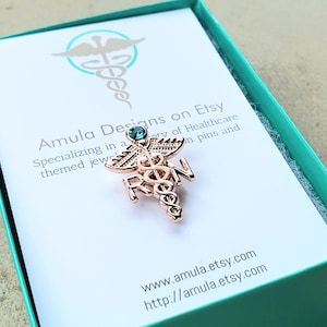 Puede incluir: Un broche de caduceo en tono oro rosa con las letras RN y un peque&ntilde;o cristal azul en la parte superior. El broche se muestra sobre una tarjeta blanca dentro de una caja color cerceta. La tarjeta incluye el texto Amula Designs on Etsy.