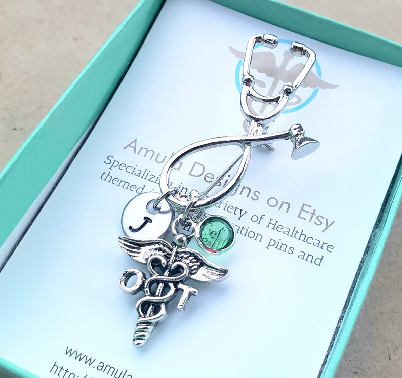 May include: Silver stethoscope charm with a green crystal, a silver medical caduceus charm with the letters "OT" and a silver initial charm with the letter "J". The charm is in a teal gift box with a white card that reads "Amula Designs on Etsy Specializing in a variety of Healthcare themed graduation pins and www.amula htto"