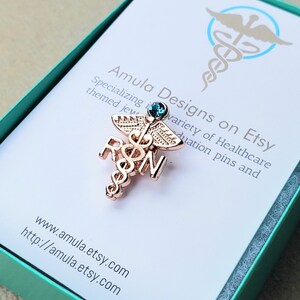 Puede incluir: Un broche de metal en tono oro rosa que presenta un s&iacute;mbolo de caduceo con las letras RSN integradas en el dise&ntilde;o. El broche est&aacute; rematado con un peque&ntilde;o cristal redondo de color azul oscuro. Se muestra sobre una tarjeta blanca con el texto Amula Designs on Etsy.