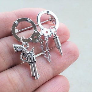 May include: Silver-toned brooch featuring a pair of handcuffs with a chain and a small gun charm. The brooch is designed to look like a pair of handcuffs with a chain attached to a small gun charm.