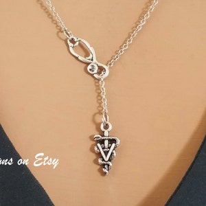 May include: Silver necklace with a stethoscope charm and a caduceus pendant. The necklace is on a model wearing a black shirt.