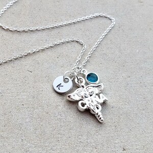May include: Silver necklace with a charm of a caduceus, a small blue gemstone, and a silver disc with the letter K.