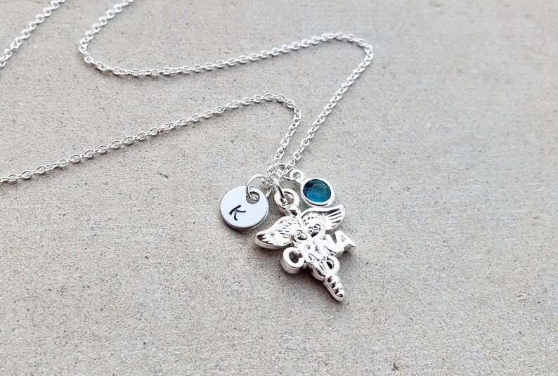 May include: Silver necklace with a charm of a caduceus, a small blue gemstone, and a silver disc with the letter K.