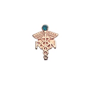 Puede incluir: Un broche de metal en tono oro rosa que presenta el s&iacute;mbolo m&eacute;dico del caduceo con las letras R y N integradas en el dise&ntilde;o. La parte superior del bast&oacute;n est&aacute; adornada con un &uacute;nico estr&aacute;s redondo de color cerceta engastado en un marco circular.