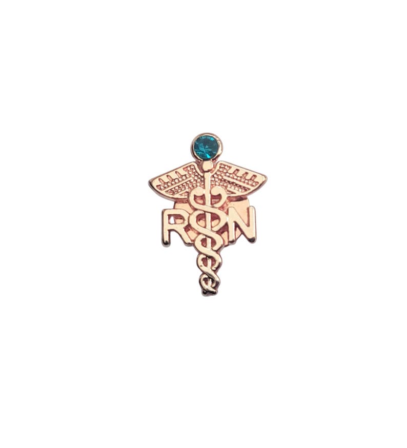 Puede incluir: Un broche de metal en tono oro rosa que presenta el s&iacute;mbolo m&eacute;dico del caduceo con las letras R y N integradas en el dise&ntilde;o. La parte superior del bast&oacute;n est&aacute; adornada con un &uacute;nico estr&aacute;s redondo de color cerceta engastado en un marco circular.