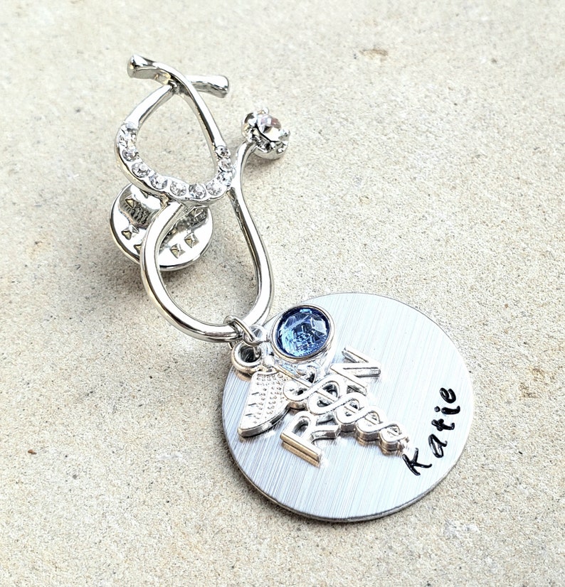 Op de afbeelding: Zilveren stethoscoopbedel met een blauwe strasssteen en een zilveren bedel met een caduceussymbool en de naam "Katie" gegraveerd op een zilveren cirkel.