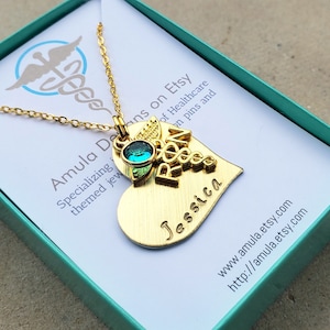 Puede incluir: Collar con colgante de corazón de oro, adornado con un amuleto de símbolo médico y la palabra "RN". El colgante también tiene el nombre "Jessica" grabado. El collar está en una caja de regalo turquesa con el nombre de la tienda Etsy "Amula Deans" impreso en ella.
