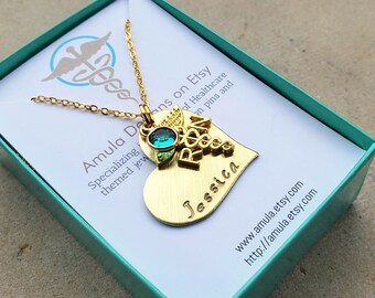 May include: Gold heart-shaped pendant necklace with a medical symbol charm and the word "RN" on it. The pendant also has the name "Jessica" engraved on it. The necklace is in a teal gift box with the Etsy shop name "Amula Deans" printed on it.