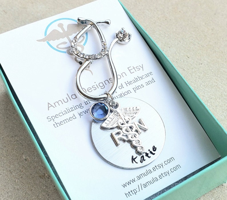 Op de afbeelding: Zilveren stethoscoopbedel met strass en een blauwe kristal. De bedel is bevestigd aan een ronde zilveren hanger met de letters "RN" en een caduceussymbool. De hanger is gegraveerd met de naam "Katie".