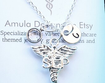 May include: Silver necklace with a medical caduceus charm, a clear crystal charm, and a silver charm with the letter "J". The necklace is displayed on a white card with the text "Amula Designs Etsy" and website information.