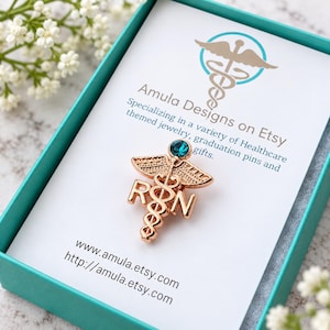 Puede incluir: Un broche de caduceo RN en tono oro rosa con una peque&ntilde;a piedra preciosa circular de color verde azulado en la parte superior. El broche se presenta sobre una tarjeta blanca dentro de una caja verde azulado, con el texto Amula Designs on Etsy.