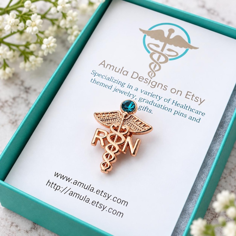 Puede incluir: Un broche de caduceo RN en tono oro rosa con una peque&ntilde;a piedra preciosa circular de color verde azulado en la parte superior. El broche se presenta sobre una tarjeta blanca dentro de una caja verde azulado, con el texto Amula Designs on Etsy.