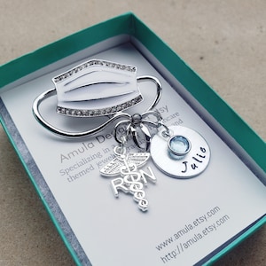 May include: A silver-toned brooch featuring a rhinestone-encrusted face mask, a charm with the letters "RN" and a caduceus, a small blue gemstone, and a round charm with the name "Julie" engraved on it. The brooch is displayed in a teal gift box.
