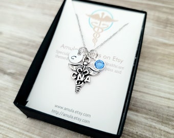 May include: A silver necklace with a charm of a caduceus with the letters CNA, a small silver disc with the letter C, and a light blue crystal charm. The necklace is in a black gift box with a white card that says "Amul-Designs on Etsy" and "Special themed healthcare pins and".