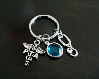 Puede incluir: Un llavero plateado con un dije de símbolo médico, un dije de cristal azul y un dije de estetoscopio.