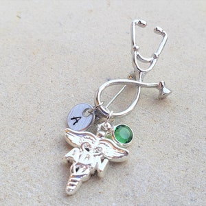 May include: A silver-toned stethoscope pin with a Caduceus charm, a green gemstone, and a small disc with the letter "A". The pin is designed to be worn and is likely a medical or nursing-themed accessory.