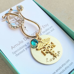 Puede incluir: Encanto de estetoscopio dorado con pedrería, un cristal verde y un encanto de símbolo médico dorado con el texto "R&N Lauren" en un disco redondo dorado.