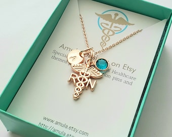 May include: A gold necklace with a medical caduceus charm, a small round charm with the letter A, and a turquoise blue crystal. The necklace is in a teal gift box with a white card that says "Amula Designs on Etsy" and "Special themed medical pins and more".