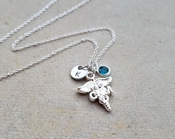 May include: Silver necklace with a medical caduceus charm, a small blue gemstone, and a personalized initial charm. The initial charm is engraved with the letter "K".