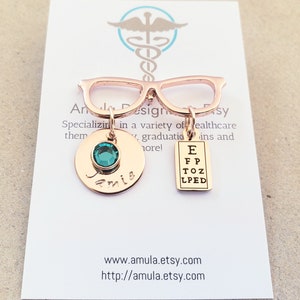 May include: Rose gold eyeglasses charm with a green gemstone, a personalized disc with the name "Jamie" and an eye chart. The charm is on a white card with the text "AnDesignsy" and a website address.