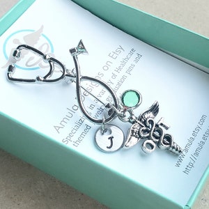 May include: Silver stethoscope charm with a green rhinestone, a silver medical caduceus charm, and a silver initial charm with the letter J. The charms are attached to a silver chain. The charms are in a small, light blue gift box with a white card that reads "Amula Designs on Etsy" and "Specializing in a variety of Healthcare themed jewelry graduation pins and www.amula.etsy.com http://amula.etsy.com".