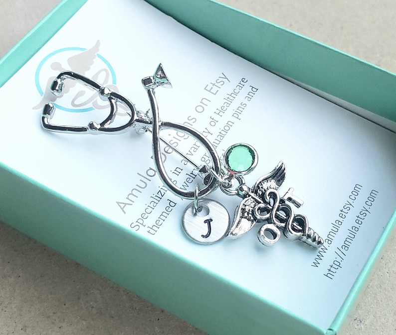 May include: Silver stethoscope charm with a green rhinestone, a silver medical caduceus charm, and a silver initial charm with the letter J. The charms are attached to a silver chain. The charms are in a small, light blue gift box with a white card that reads "Amula Designs on Etsy" and "Specializing in a variety of Healthcare themed jewelry graduation pins and www.amula.etsy.com http://amula.etsy.com".