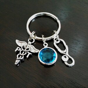 Puede incluir: Llavero plateado con tres colgantes: un símbolo médico, una gema azul redonda y un estetoscopio. Los colgantes cuelgan de un anillo plateado. El fondo es una superficie de madera oscura y pulida.
