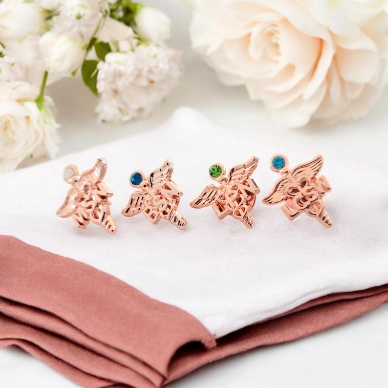 Puede incluir: Cuatro broches de metal en tono oro rosa con forma de s&iacute;mbolos de caduceo con alas. Cada broche presenta un peque&ntilde;o estr&aacute;s redondo de color en la parte superior central y letras grabadas en la parte inferior, incluyendo MSN, BSN, LPN y CSN.