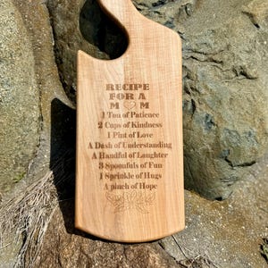 May include: A wooden chopping board with a handle, engraved with the text "RECIPE FOR A MOM" and a list of ingredients for a "Mom" including patience, kindness, love, understanding, laughter, fun, hugs, and hope.