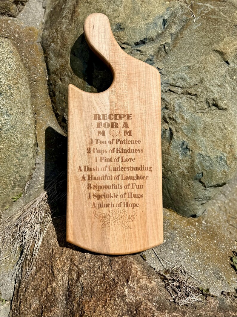 May include: A wooden chopping board with a handle, engraved with the text "RECIPE FOR A MOM" and a list of ingredients for a "Mom" including patience, kindness, love, understanding, laughter, fun, hugs, and hope.