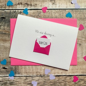 May include: A white card with the text "We are having a..." and a pink envelope with a card that reads "BABY GIRL". The card is on top of a pink envelope, with heart-shaped confetti scattered around.