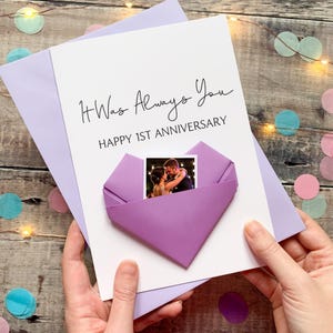 A hand holding a handmade card with a photo of a couple inside a heart-shaped origami. The card is purple and white, and the message reads "It Was Always You" and "Happy 1st Anniversary".