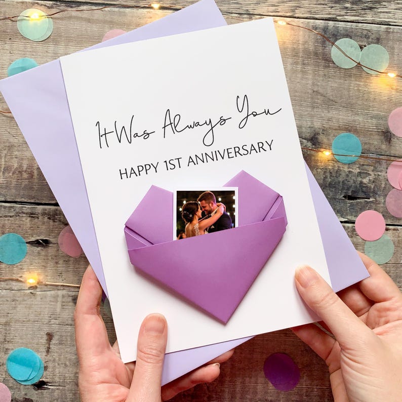 A hand holding a handmade card with a photo of a couple inside a heart-shaped origami. The card is purple and white, and the message reads "It Was Always You" and "Happy 1st Anniversary".