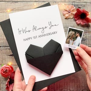 A hand holding a card with a heart-shaped origami made from black paper, surrounded by flowers and a small photograph.