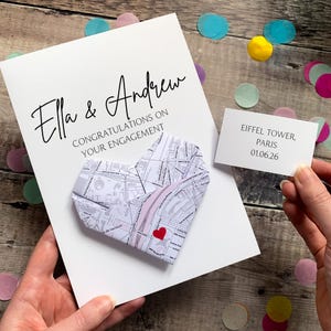 May include: A white engagement card featuring the text Ella & Andrew CONGRATULATIONS ON YOUR ENGAGEMENT with a folded paper heart made from a map of Paris. A small white card reads EIFFEL TOWER, PARIS 01.06.26. A red heart icon marks a location.