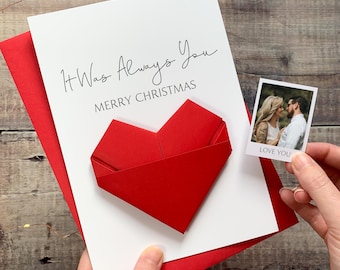 May include: A white Christmas card with the text "It Was Always You" and "Merry Christmas" with a red origami heart. A small photo with the words "Love You" is held in the hand.