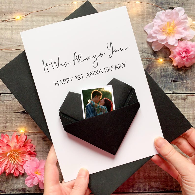 A person's hands holding a card with a photo of a couple embracing, surrounded by pink flowers and a string of lights.
