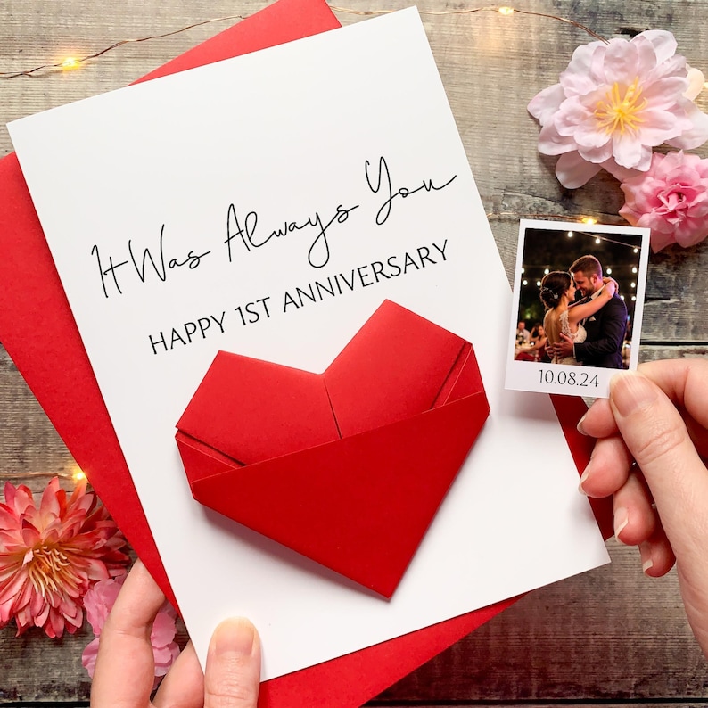 A handmade card with a red heart-shaped origami and a photograph of a couple embracing, placed on a wooden surface with pink flowers.