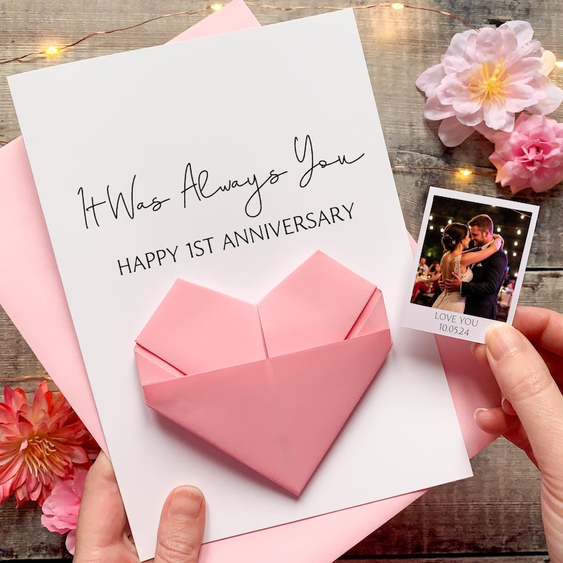 A handmade card with a pink heart-shaped origami and a photograph of a couple embracing, placed on a wooden surface with pink flowers.