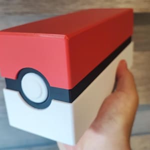 May include: A red, black, and white rectangular box with a circular white button. The box is held in a hand. The top of the box is red, the middle is black, and the bottom is white. The box is a collectible item.