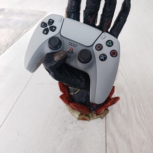 May include: A grey PlayStation 5 controller held by a black, textured hand-shaped stand with red and gold accents. The controller has black buttons and a directional pad.