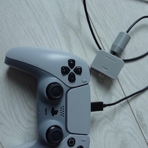 May include: A grey PlayStation 5 DualSense controller with a black USB cable connected to it. A grey Sony PlayStation adapter is connected to the other end of the cable. The controller and adapter are on a light grey wooden surface.