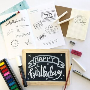 May include: A white chalk marker, a black chalkboard with the words "HAPPY BIRTHDAY" written in white chalk, a pink eraser, a white paint pen, a set of watercolor paints, and a book with project ideas for embellishments.
